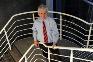 a white haired formally suited smiling man on top of a staircase